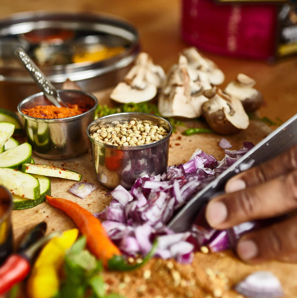 Lifestyle photo showing a hand chopping onion, other chopped vegetables and tins full of spices can also be seen.