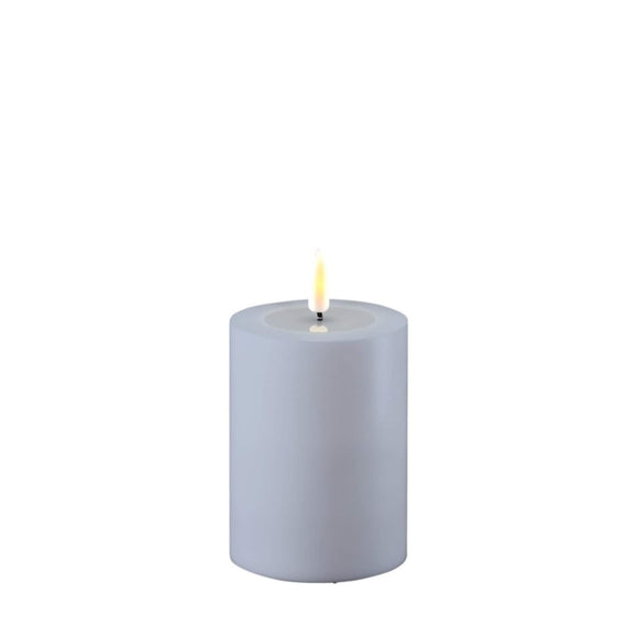 Photo of a Deluxe HomeArt dust blue LED Pillar candle that is 7.5cm wide and 10cm tall. At the top of the candle is a clear resign pool that looks like a pool of melted wax. The flame is white and orange and is on a black wick.