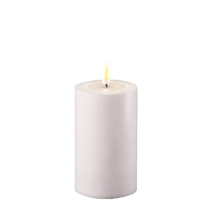 Photo of a Deluxe HomeArt white outdoor LED Pillar candle that is 7.5cm wide and 12.5cm tall. At the top of the candle is a clear resign pool that looks like a pool of melted wax. The flame is white and orange and is on a black wick.