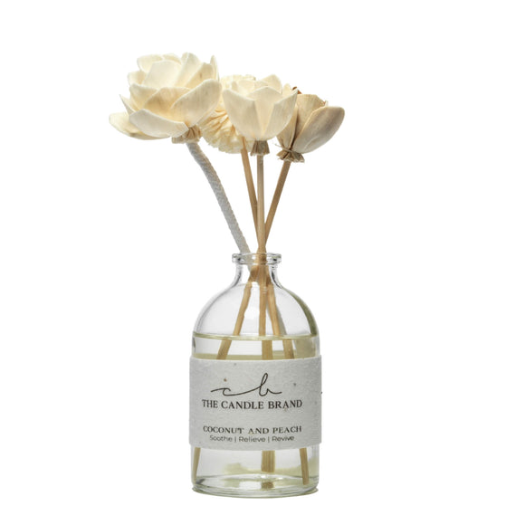 Photo of a clear glass bottle that is 3/4 full of diffuser oil. Four beige flowers on reeds and a cotton wick tree in the jar like a mini bouquet. The bottle has a hand made paper label wrapped around it. Some of the embedded flowers seeds can be seen in the label. The Candle Brand logo can be seen on the top half of the label front. Below this is the words ‘Coconut and Peach. Soothe | Relieve | Revive’.