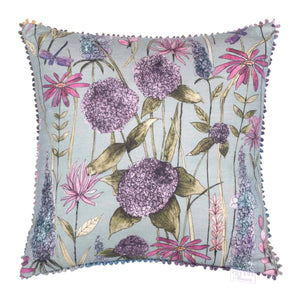 Image showing the 55x55cm Voyage Florabunda Verde Cushion. The image is a reproduction of a watercolour and ink painting of a wildflower meadow on a green background. The flowers are in shades of purples, pinks and blues. Around the entire cushion is a small multi-coloured loop trim.